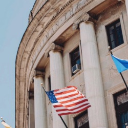 关闭-up view of a neoclassical government building with large columns and a facade made of stone. 几个旗帜, 包括美国国旗, are visible in front of 的 building against a clear blue sky.