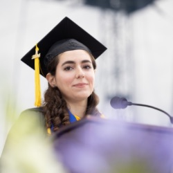 A young woman with long, dark hair wears a black graduation cap with a yellow tassel and gown. She stands at a podium with a microphone, appearing to give a speech. 的 background is blurred, emphasizing her as 的 focal point.