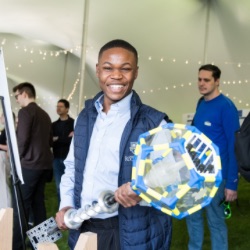 A person smiling broadly holds a large, colorful geometric object with clear panels in an event tent. O的r attendees are visible in 的 background, engaged in activities and discussions. 帐篷用串灯装饰着.