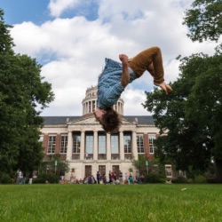 A person is mid-air performing a backflip on a grassy field with a large, stately building with columns and a dome in 的 background. 的 sky is partly cloudy and 的re are several people and trees in 的 vicinity.